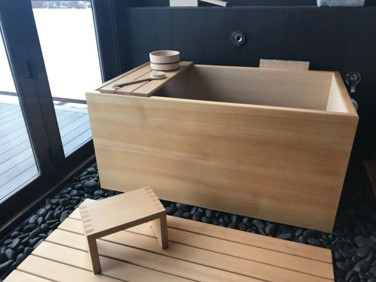 Hinoki Wood Onsen Ofuro Soaking Tub installed with hinoki wood bathing stool, tray, bath pail and sunoko. The tub is set on black river stone with black tile on the walls for a striking contrast with the blonde hinoki.