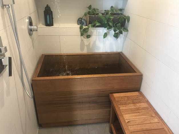 A small red cedar onsen in a white tiled alcove is filled from a waterfall faucet installed near the ceiling.